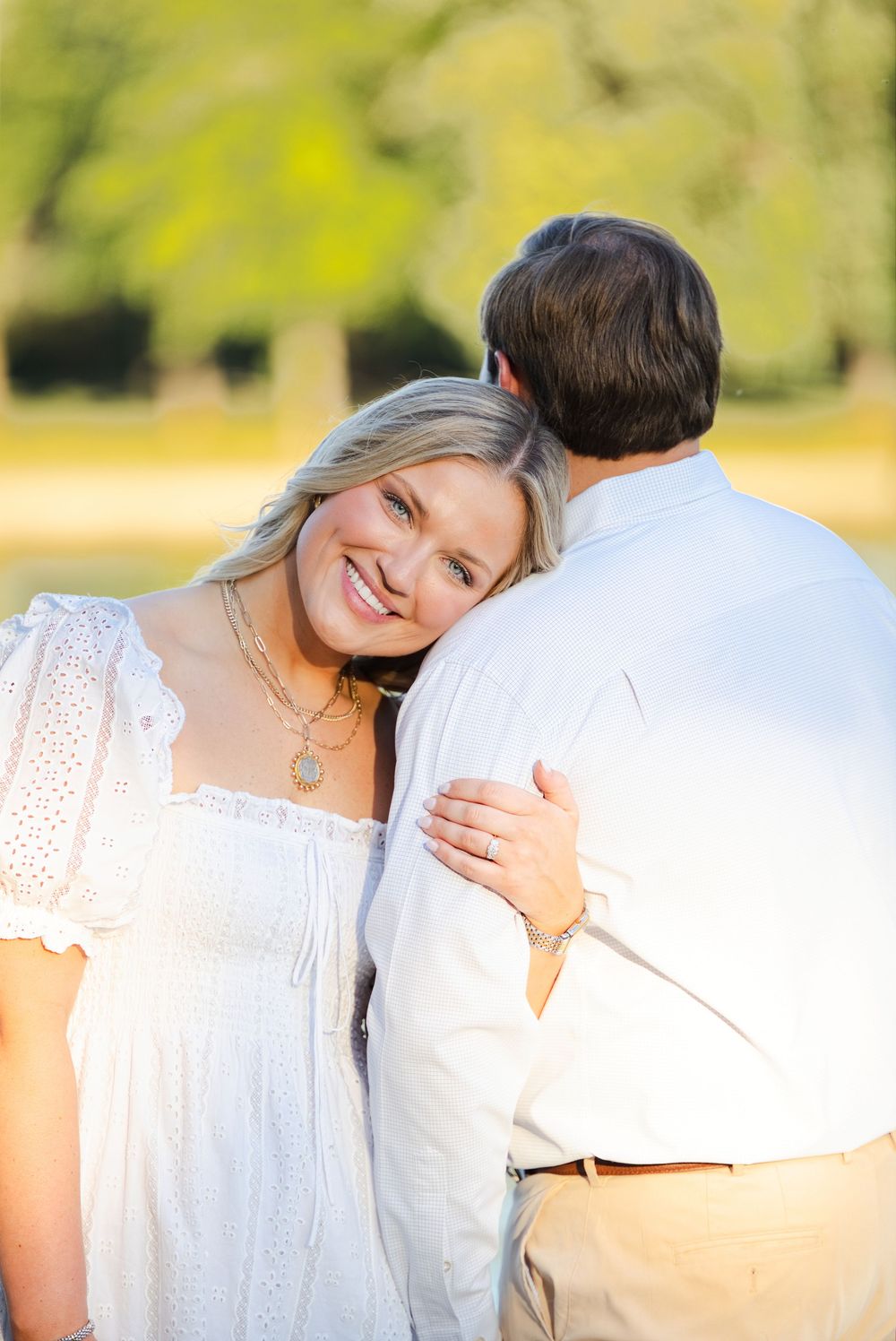 Smiling blonde woman in white dress hugs a man outdoors, leaning her head on his shoulder in a sunny park setting.