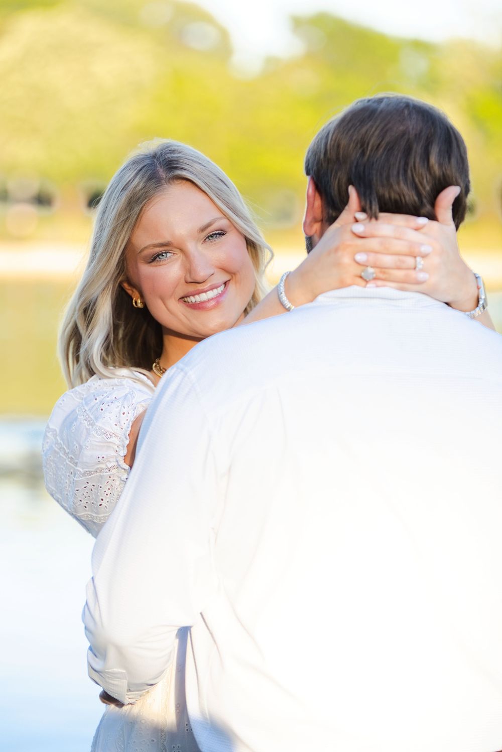 Smiling blonde woman embracing a man outdoors near a lake, wearing an engagement ring on her hand.