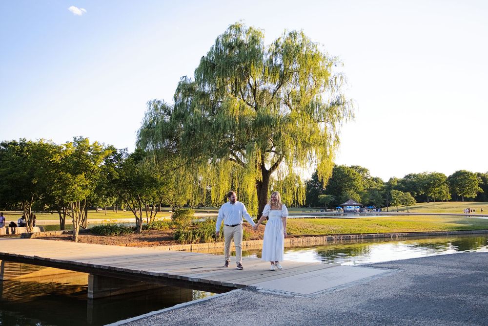 Couple holds hands on a wooden bridge near a weeping willow tree in a sunny park setting.