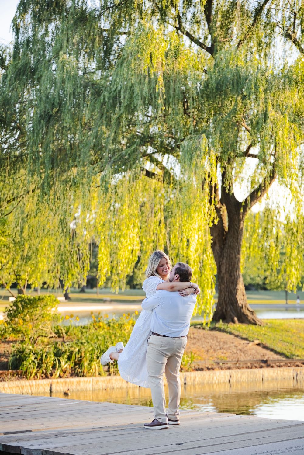 Couple embracing by a serene pond under a weeping willow tree on a wooden dock during golden hour.