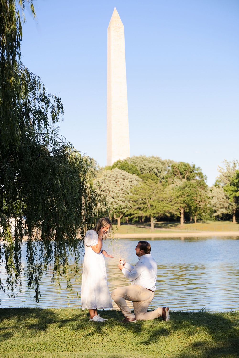 Man proposes to woman near Reflecting Pool with Washington Monument in background on sunny day.