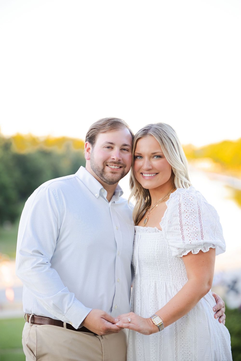 Smiling couple in white outfits holding hands outdoors at golden hour near a scenic waterway.