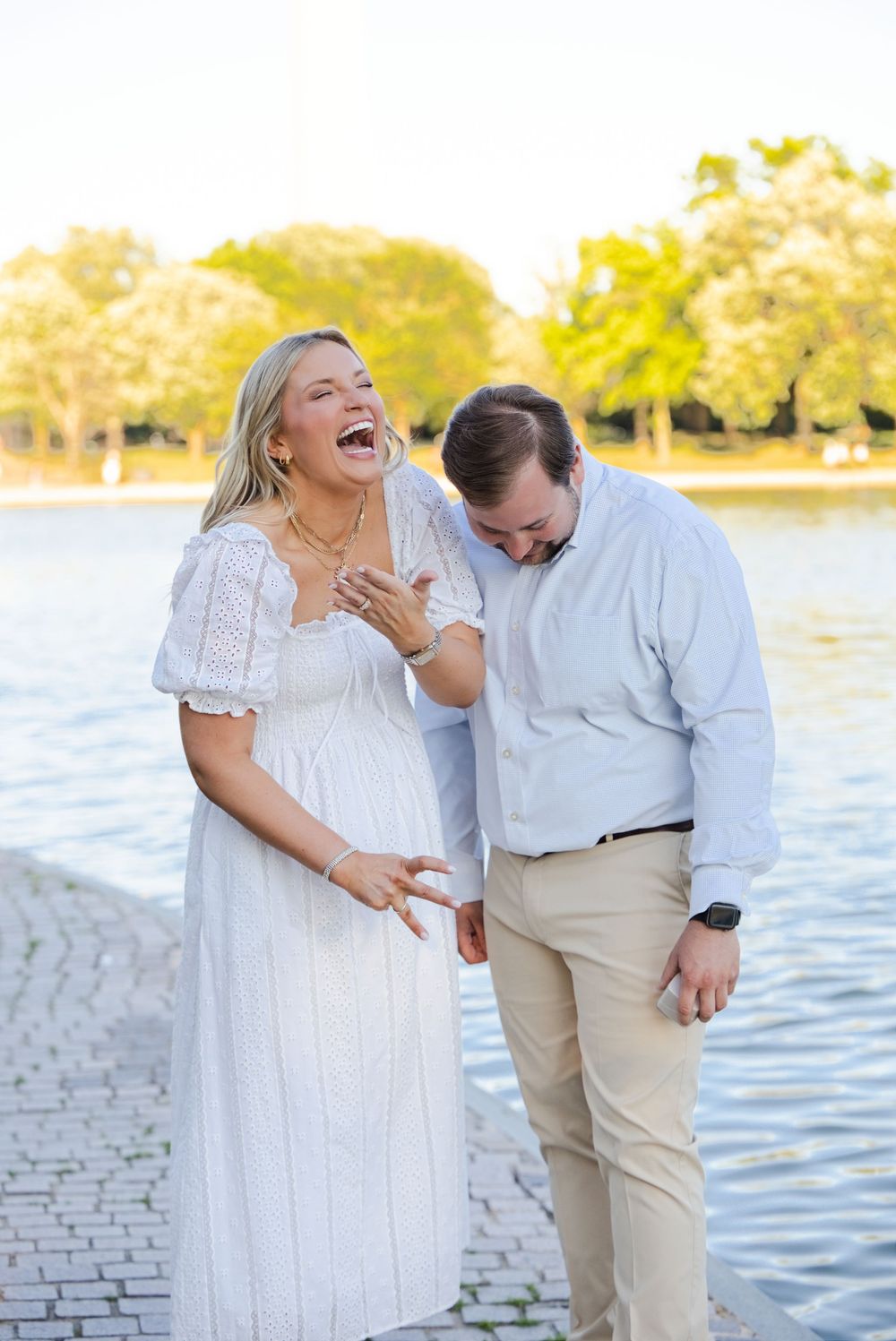 Couple laughing joyfully together by a sunlit waterfront, woman in white dress, man in light blue shirt and khaki pants.