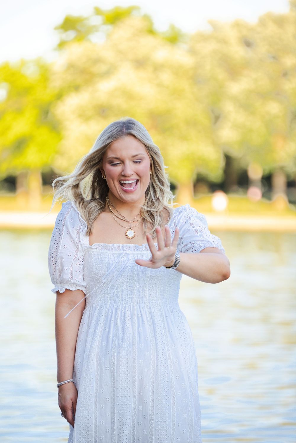 Excited blonde woman in white dress shows off engagement ring outdoors near a pond.