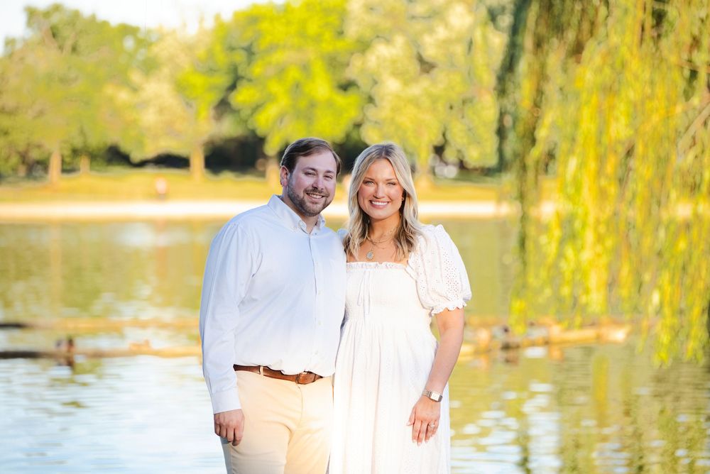 Smiling couple posing outdoors near a serene pond with lush green willow trees in a sunny park setting.