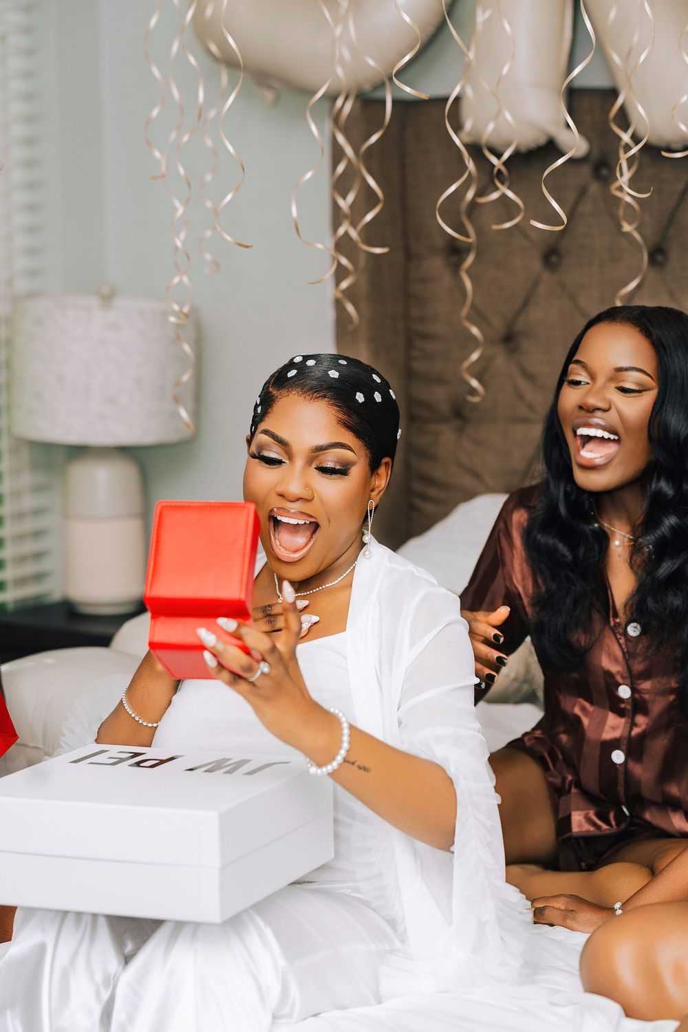 Two women in silk robes laugh joyfully while opening a red gift box during a bridal party celebration with gold balloons.