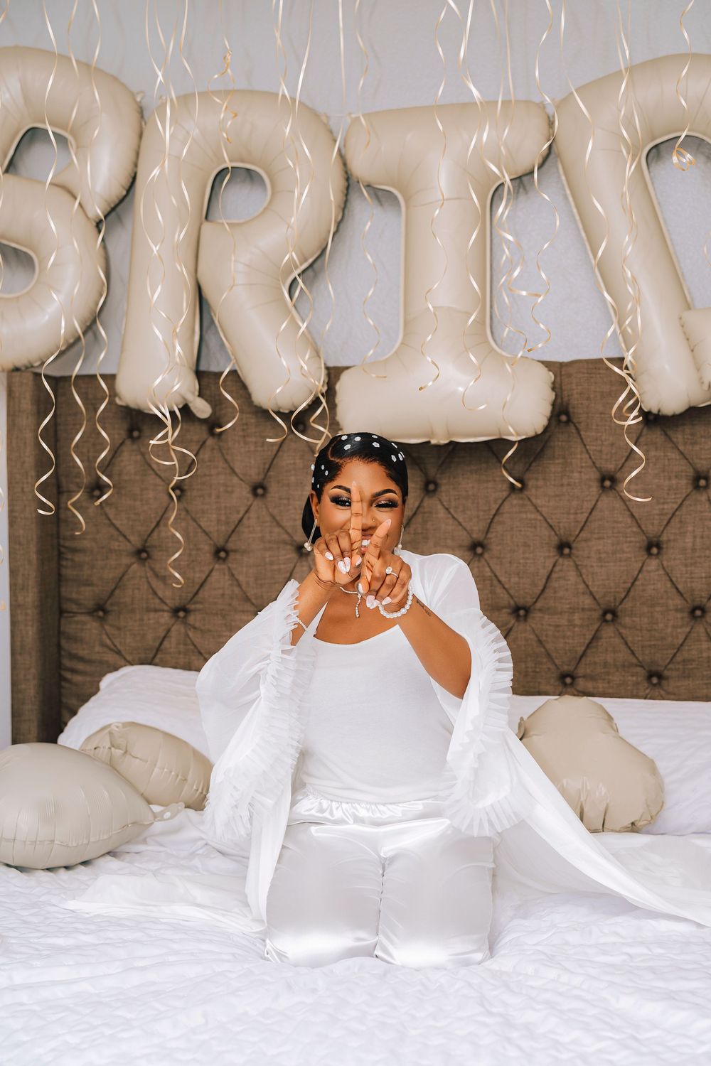 A joyful bride in white blows confetti on bed with BRIDE balloon letters and streamers.