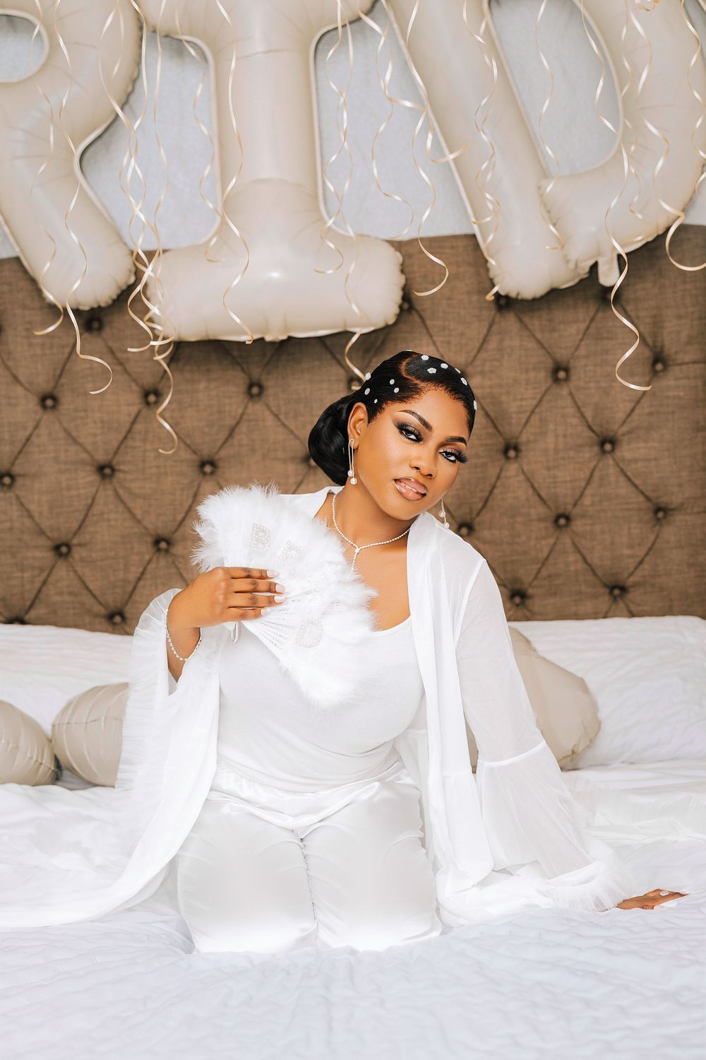 A bride in white outfit sits on bed with BRIDE balloon letters and streamers behind her.