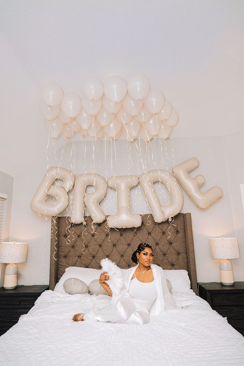 Bride sitting on white bed surrounded by white balloons and large letter balloons spelling BRIDE above tufted headboard.