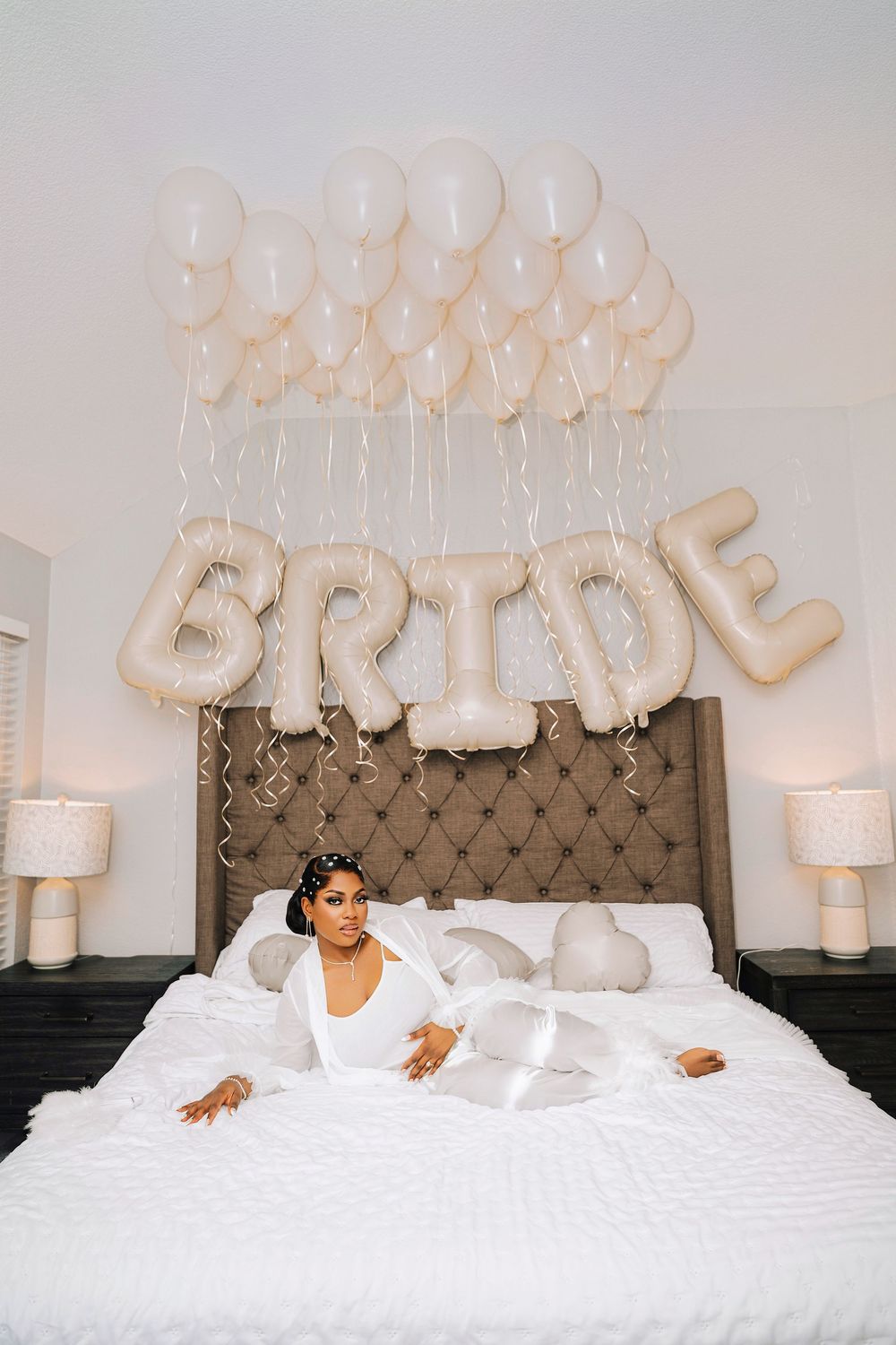 Bride in white robe poses alone on white bed beneath large BRIDE letter balloons and cream balloon ceiling display.