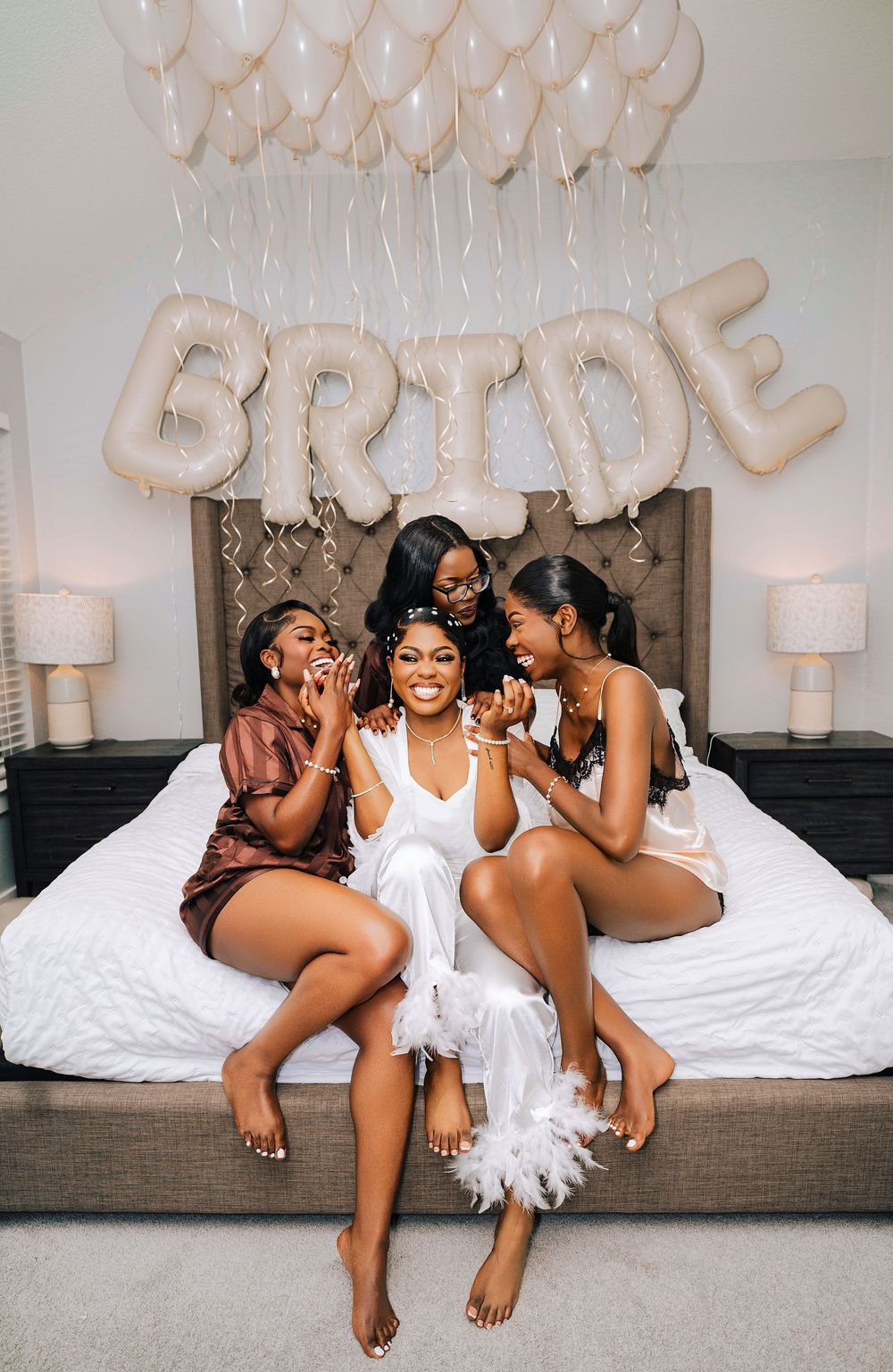 Bride in white feather robe sits on bed with three bridesmaids under BRIDE balloon letters and white balloons.