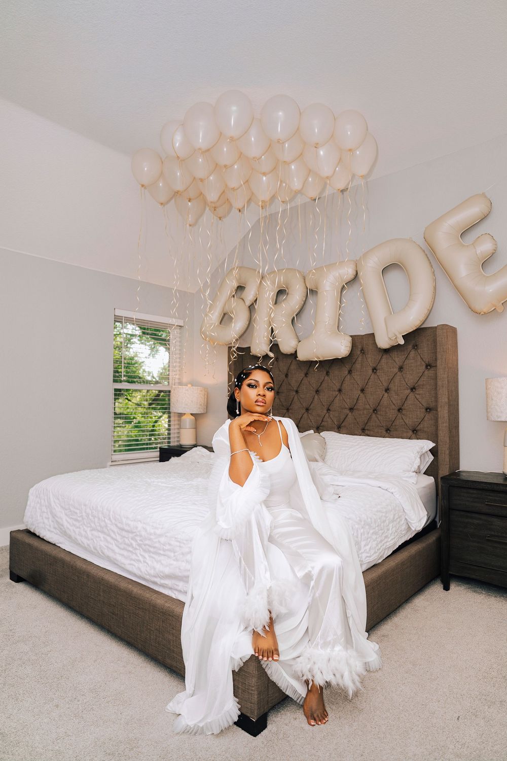Bride in white feather robe sits on bed with cream balloons and BRIDE letter balloons in decorated bedroom.