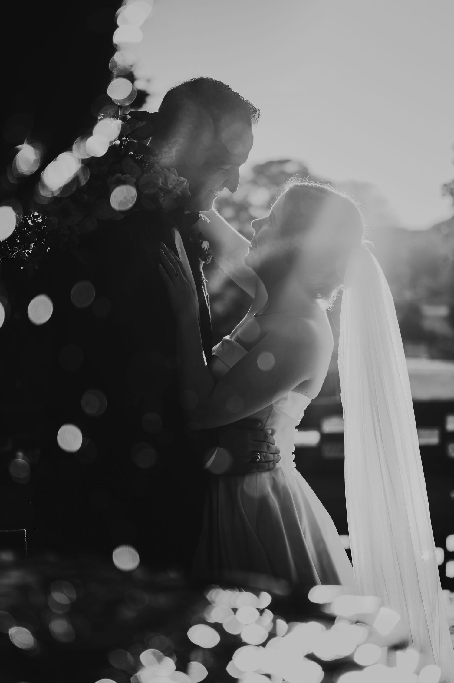 Black and white wedding photo of bride and groom embracing with bokeh lights, veil flowing, backlit by warm sunlight.
