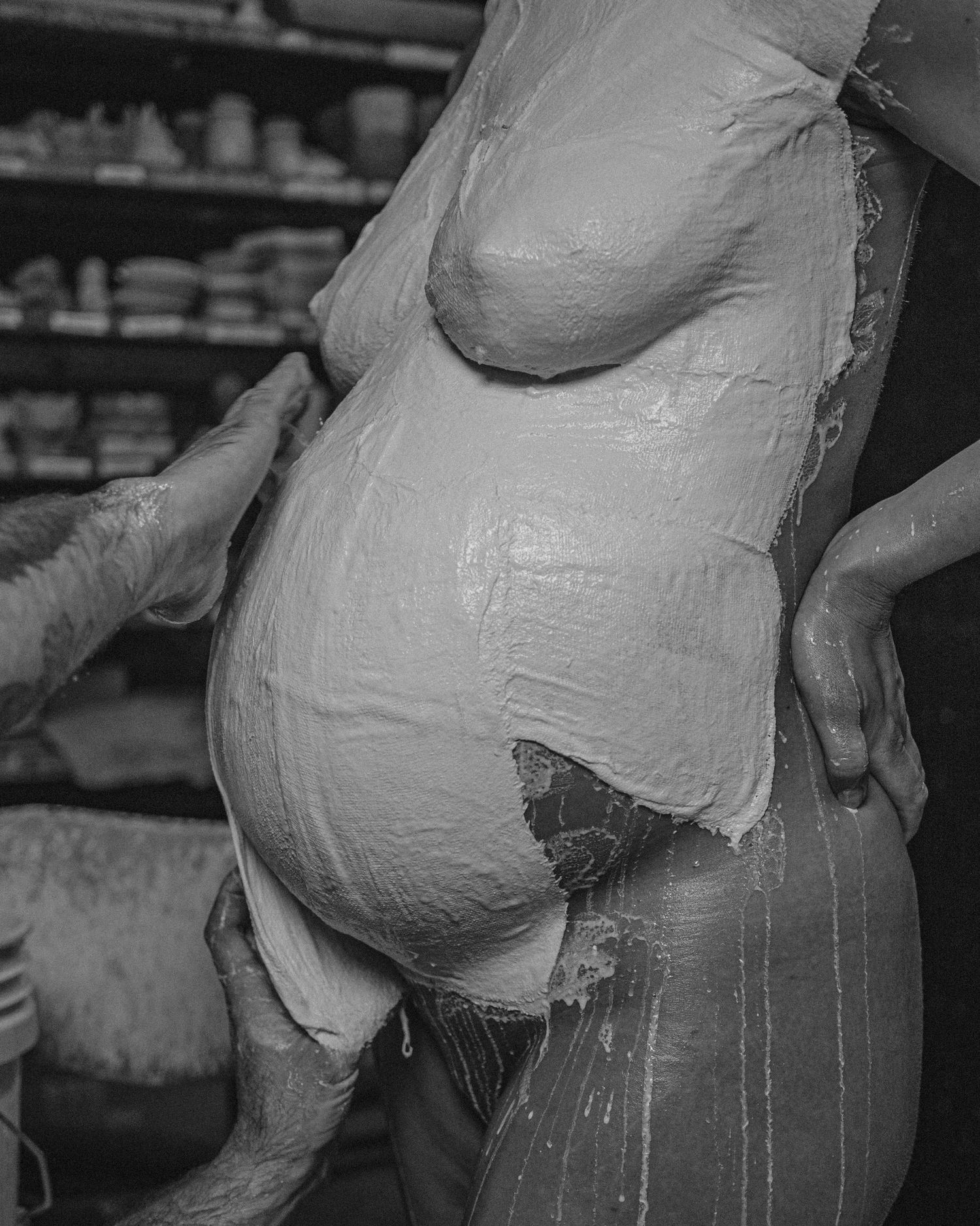 Black and white photo of a pregnant woman covered in wet clay during a body casting process in an art studio.