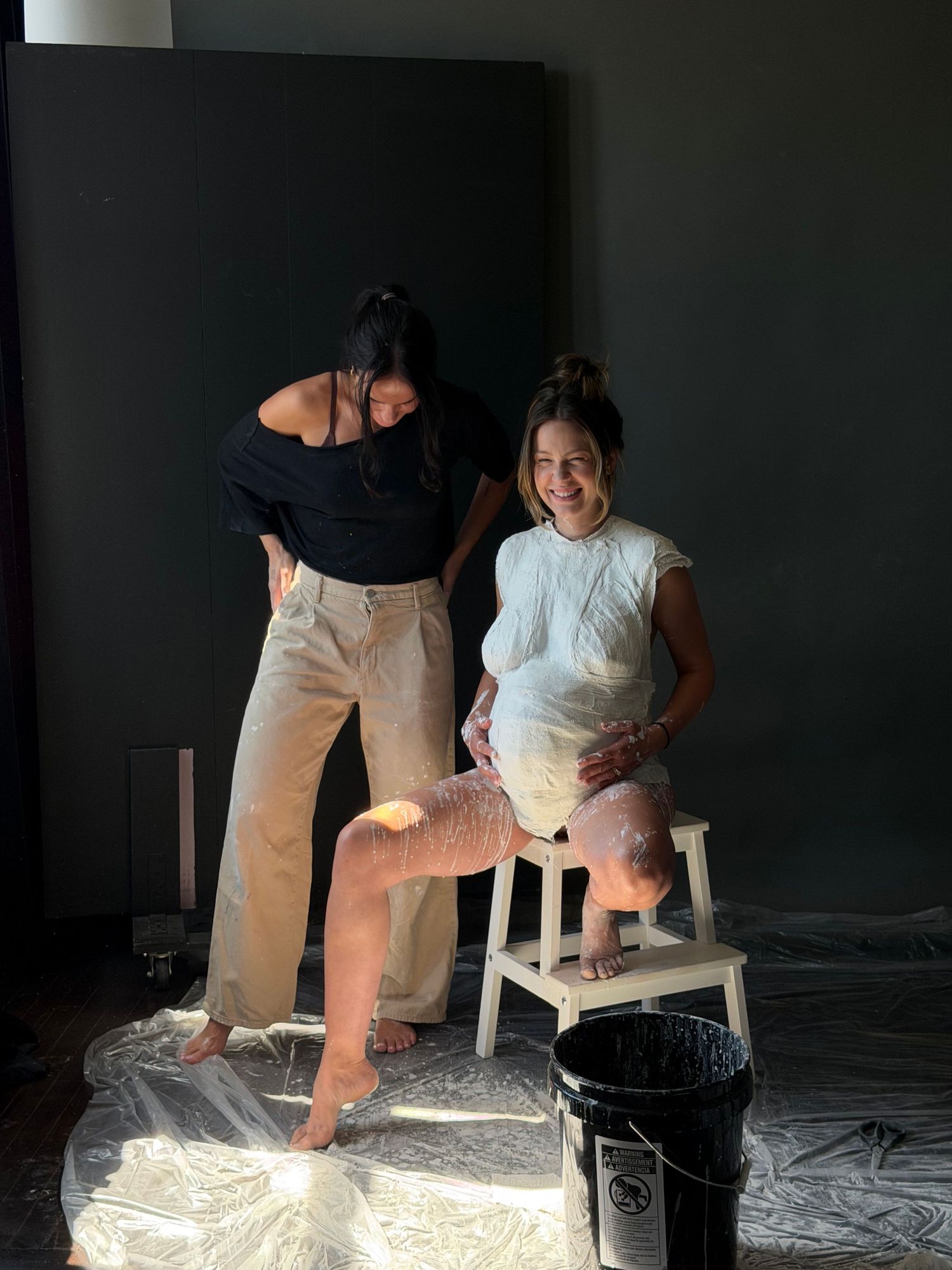 Two women in a studio setting, one pregnant getting a belly cast made, surrounded by plaster materials.