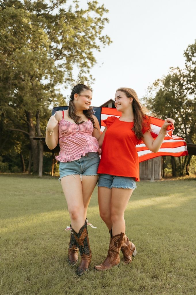 Two young women in cowboy boots and denim shorts hold an American flag outdoors on a sunny day.