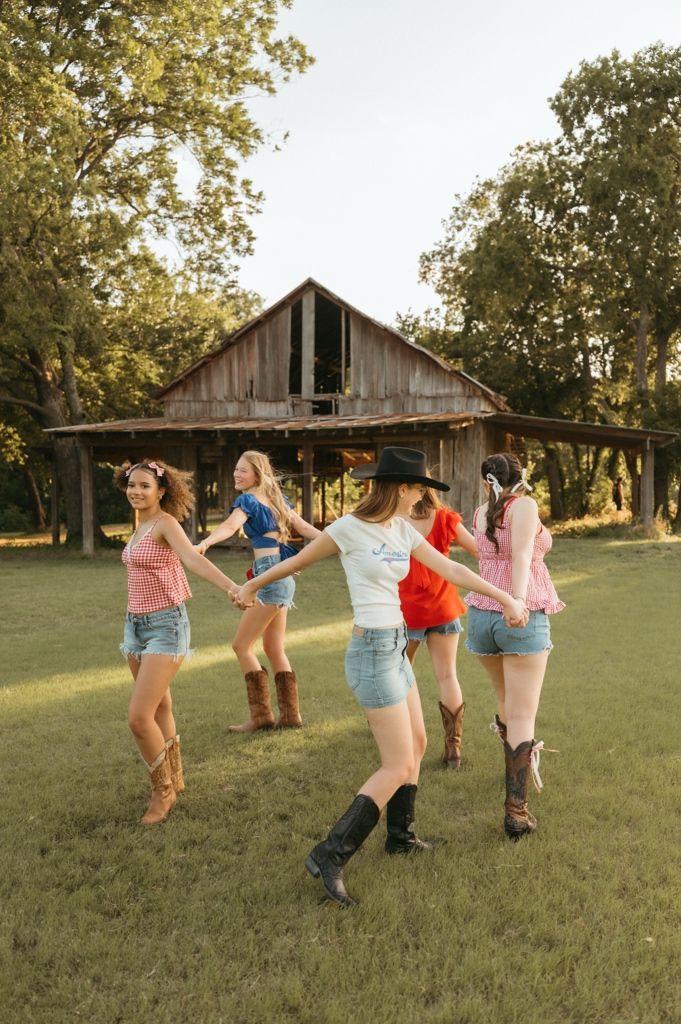 Five women in cowboy boots and denim shorts hold hands dancing in a circle on a green lawn near a rustic barn.