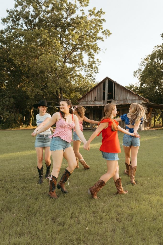Four girls in denim shorts and cowboy boots dance together in a field near a rustic barn on a sunny day.