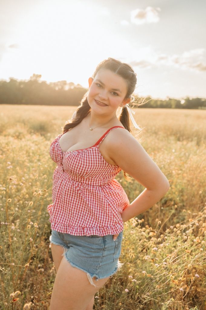 Young woman in red gingham top and denim shorts posing in a sunlit field of wildflowers.