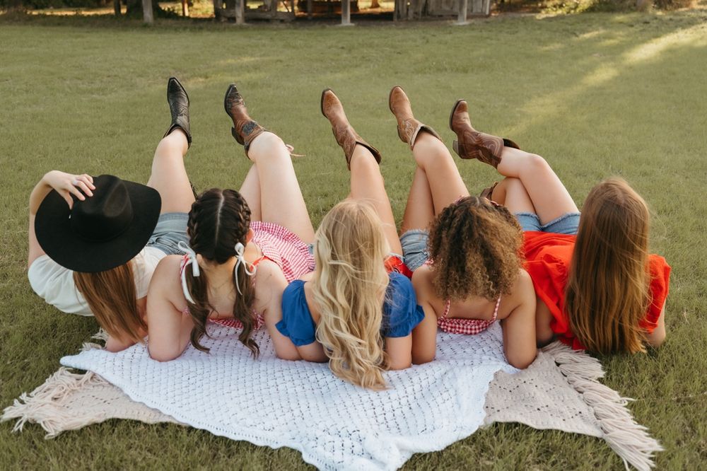 Six girls lying on a blanket in grass, legs raised, wearing denim shorts and cowboy boots, viewed from behind.