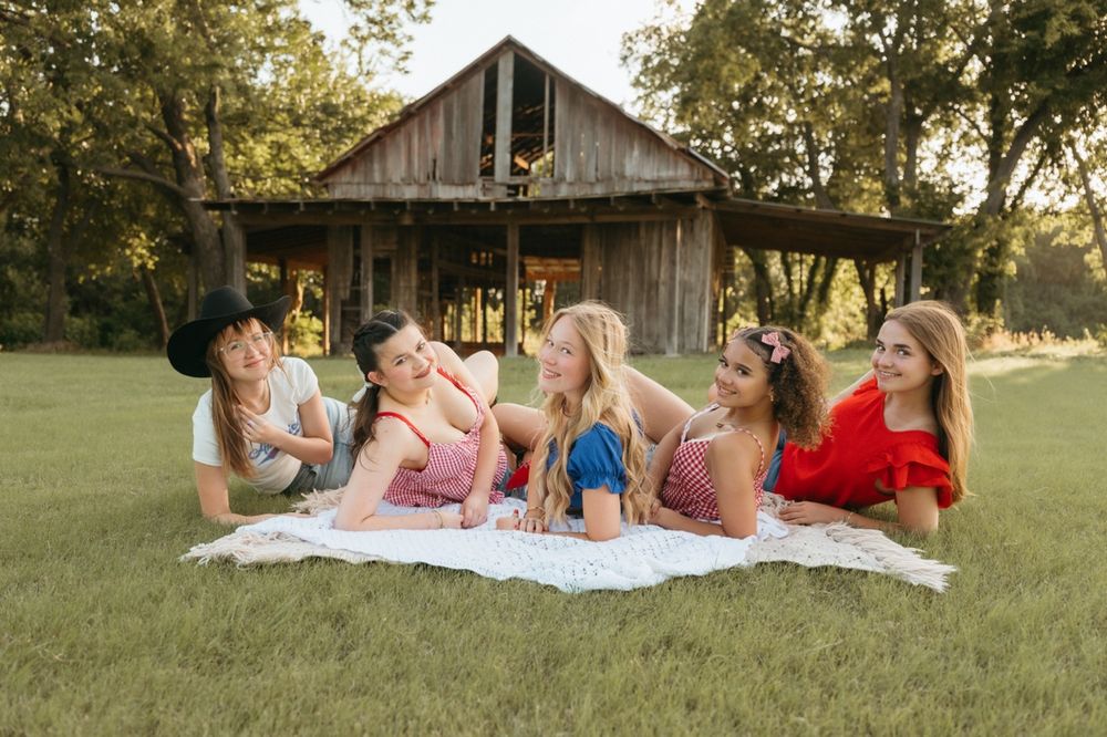 Six young women lying on a white blanket in a grassy field in front of a rustic wooden barn.