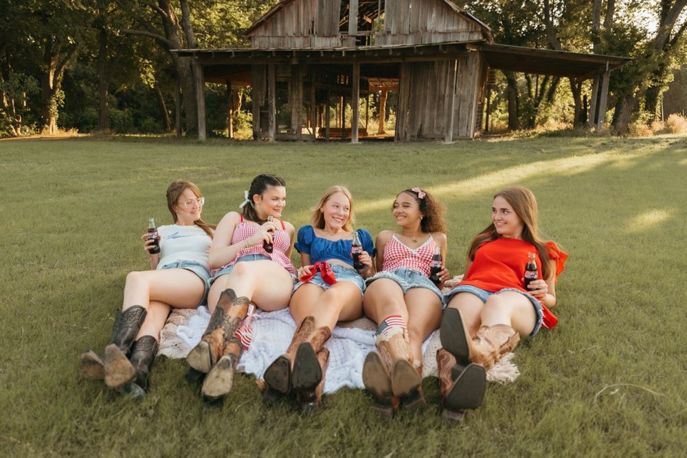 Five young women laughing and relaxing on grass in front of a rustic barn, wearing casual summer outfits.