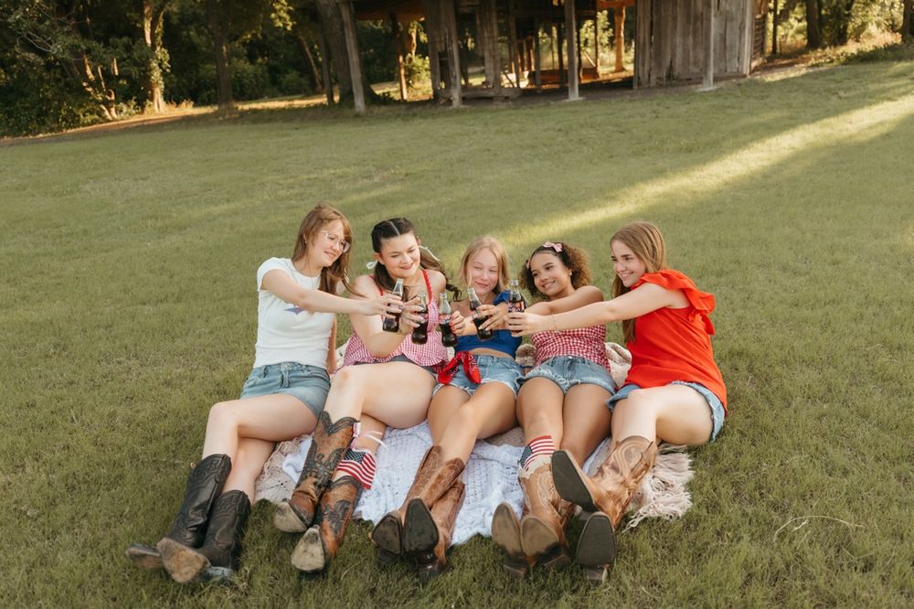 Group of girls toasting with bottles while sitting on grass in cowboy boots.