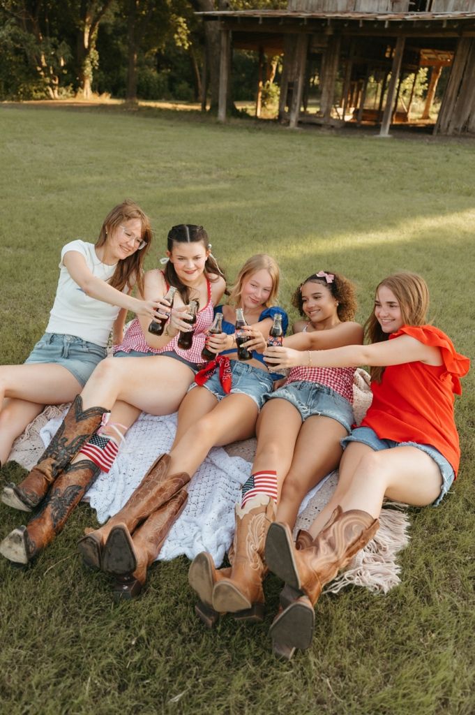 Five girls sitting on a blanket outdoors in denim shorts and cowboy boots, toasting drinks together on a sunny day.