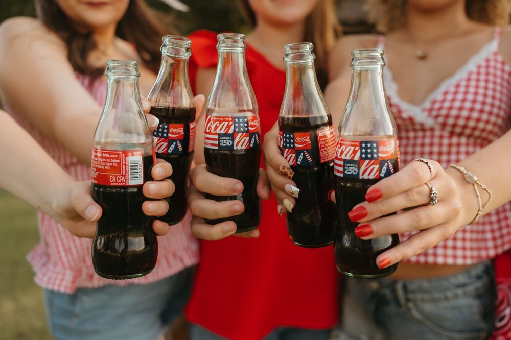 Four women clinking Coca-Cola glass bottles together in a celebratory toast outdoors.