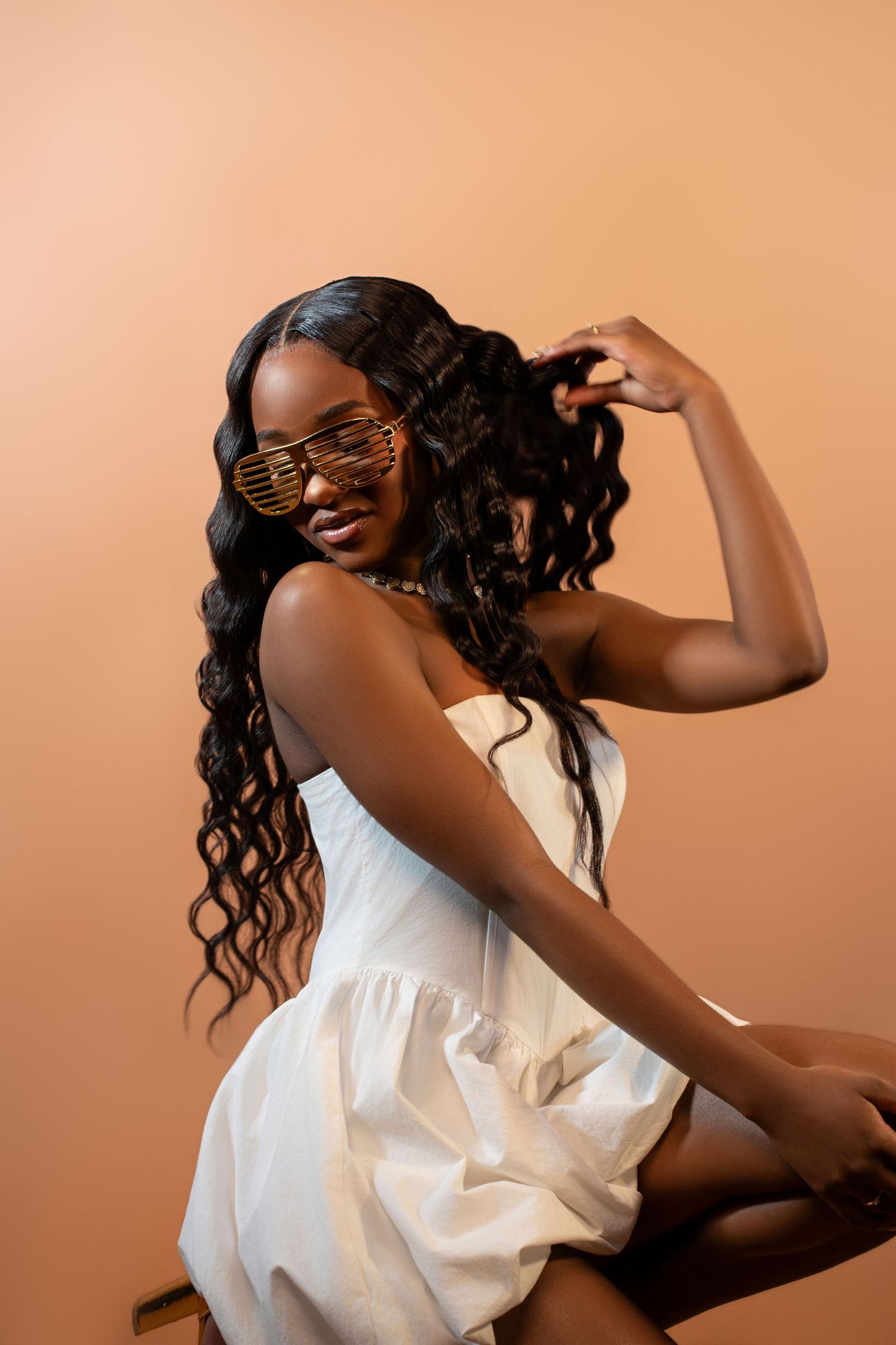 Young Black woman in white dress and gold shutter shades poses confidently against a warm beige background.