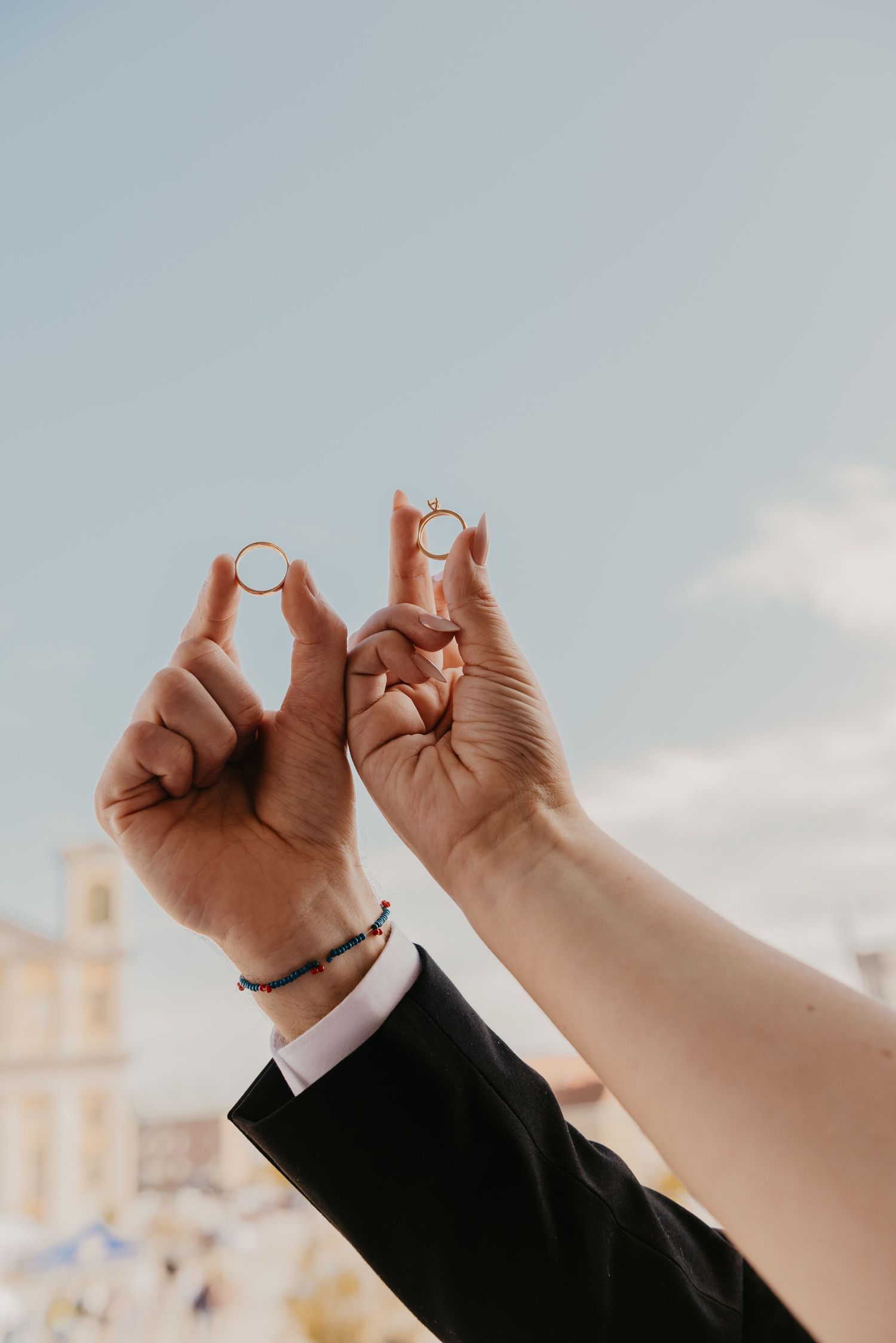 Bride and groom holding up matching gold wedding rings against a blue sky on their wedding day.