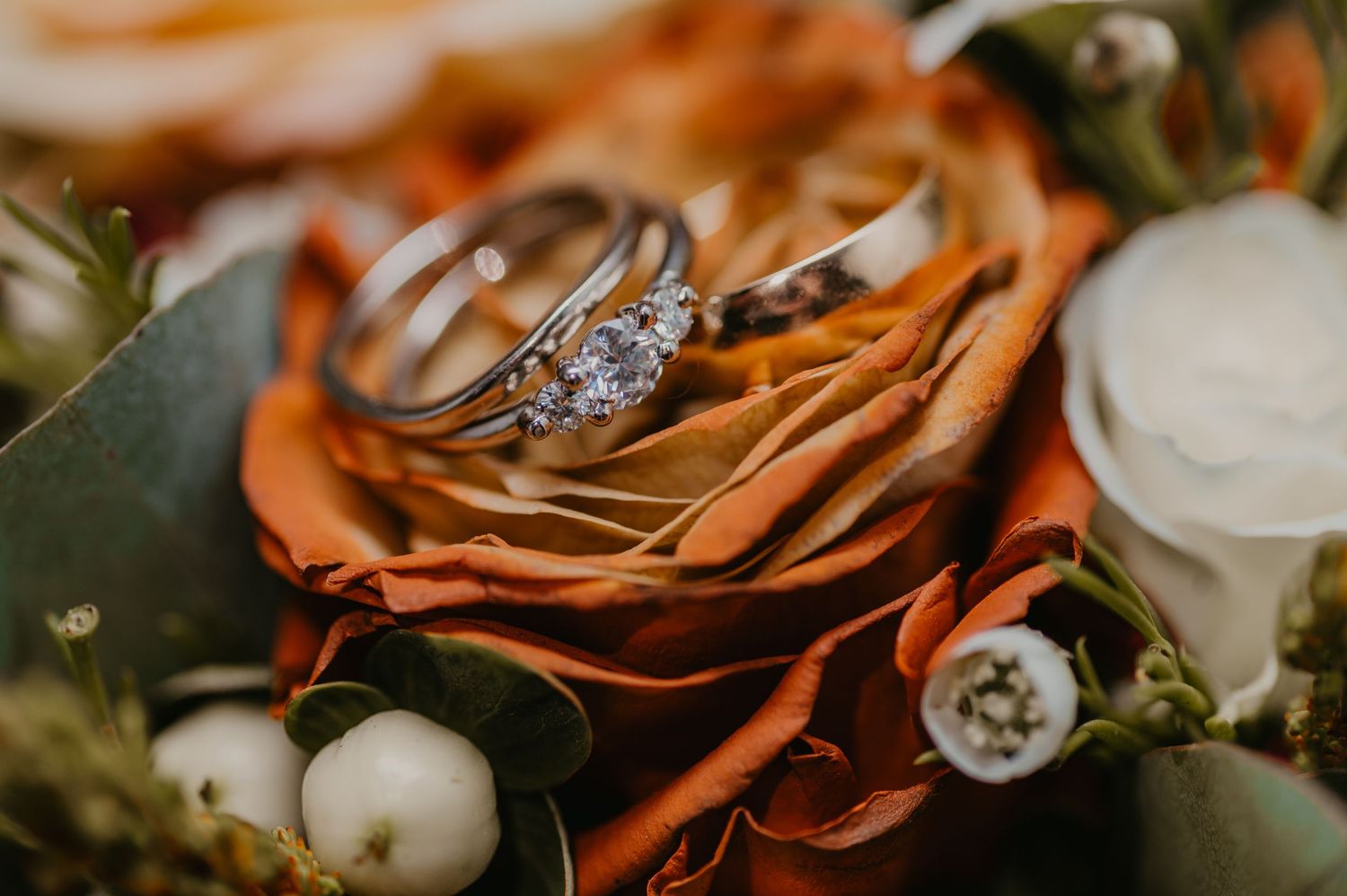 Diamond engagement ring and wedding band resting on an orange rose with white berries in a bridal bouquet.