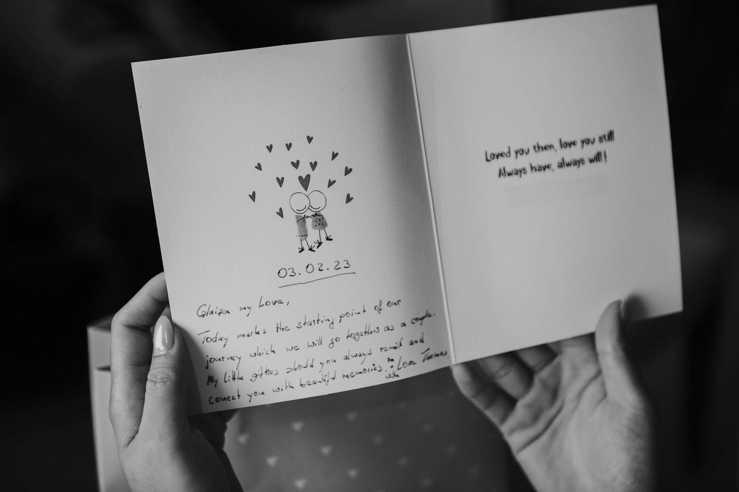 Black and white photo of hands holding open a wedding greeting card with handwritten text and a small couple illustration , on a bridal prep