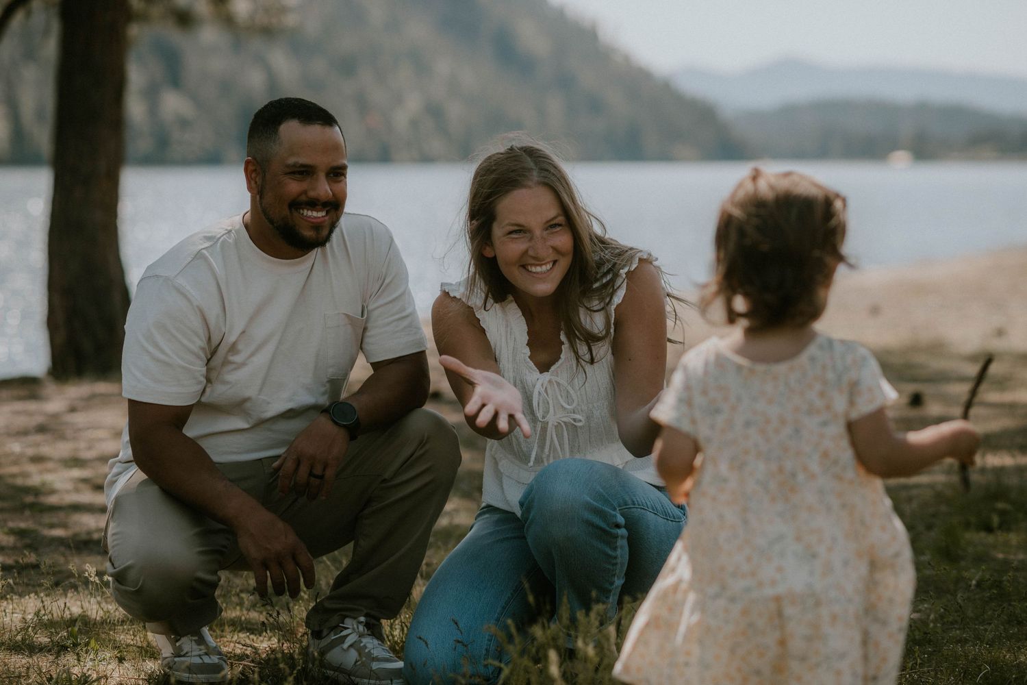 A couple smiles at their toddler daughter playing outdoors near a lake surrounded by trees.