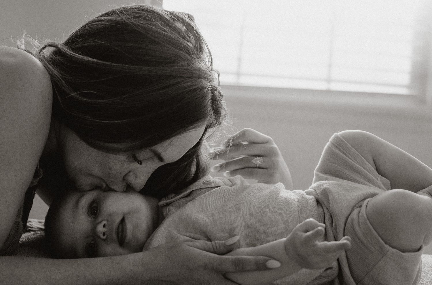 Black and white photo of a mother tenderly kissing her newborn baby while lying together in soft natural light.