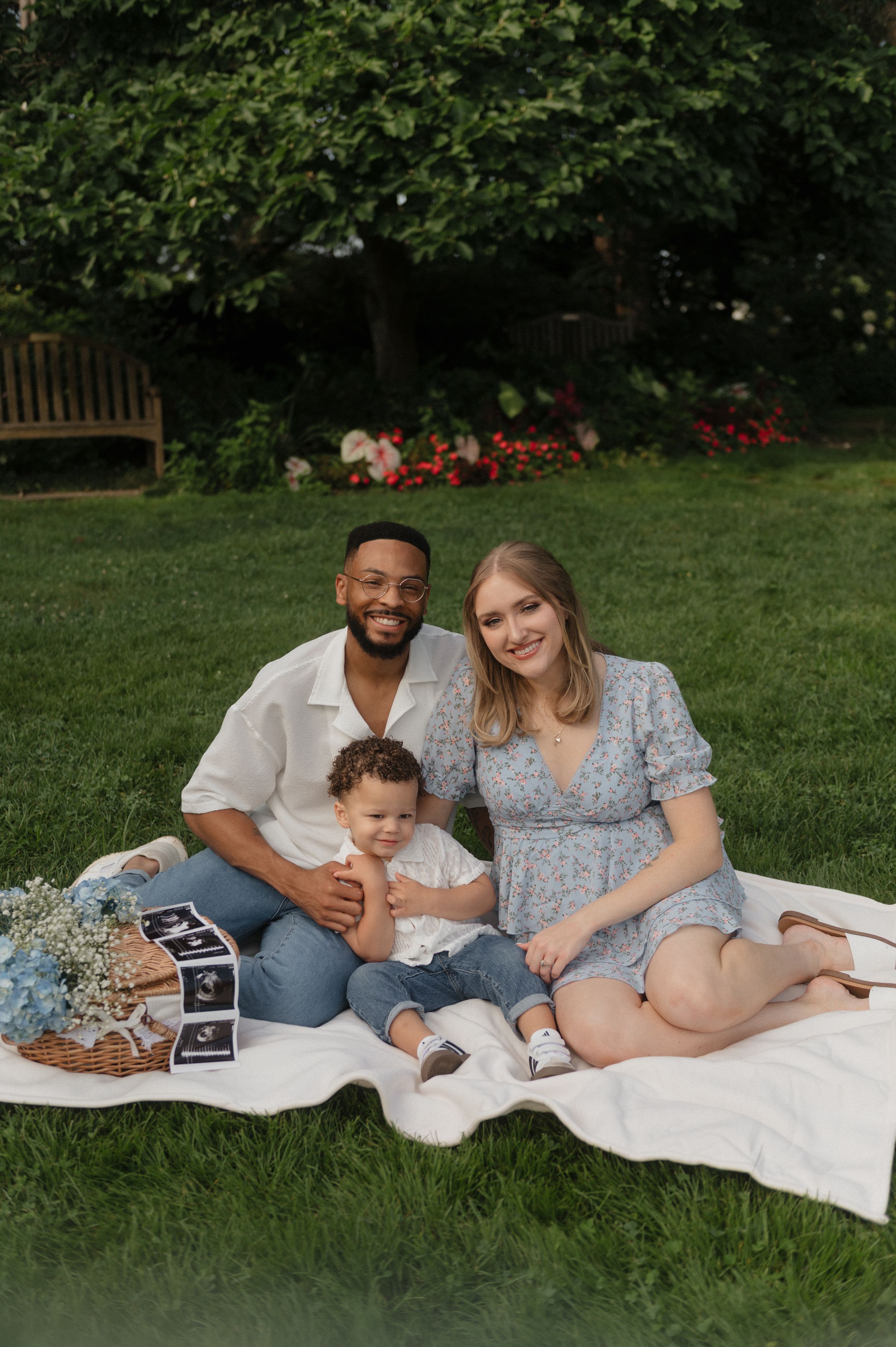 A family relaxes on a blanket in a garden with red flowers and a wooden bench visible in the background.