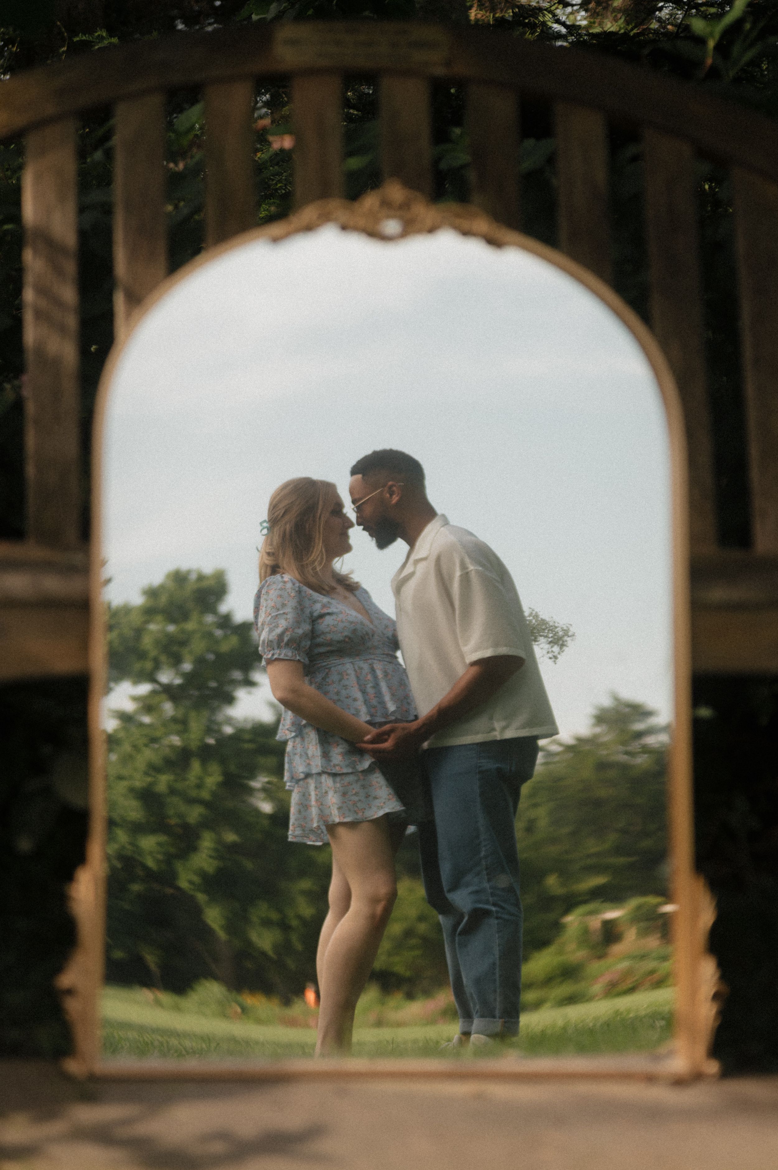 A couple shares an intimate moment beneath a wooden archway in a garden while expecting a baby.