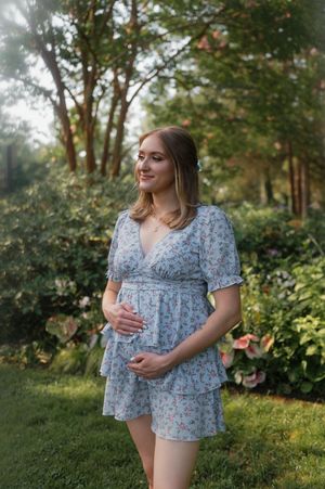 Maternity photoshoot in a garden shows woman in floral blue dress under golden sunlight during sunset.