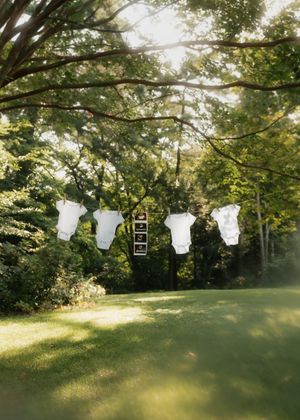 Baby clothes and onesies hanging on a clothesline outdoors against a sunlit green garden background.