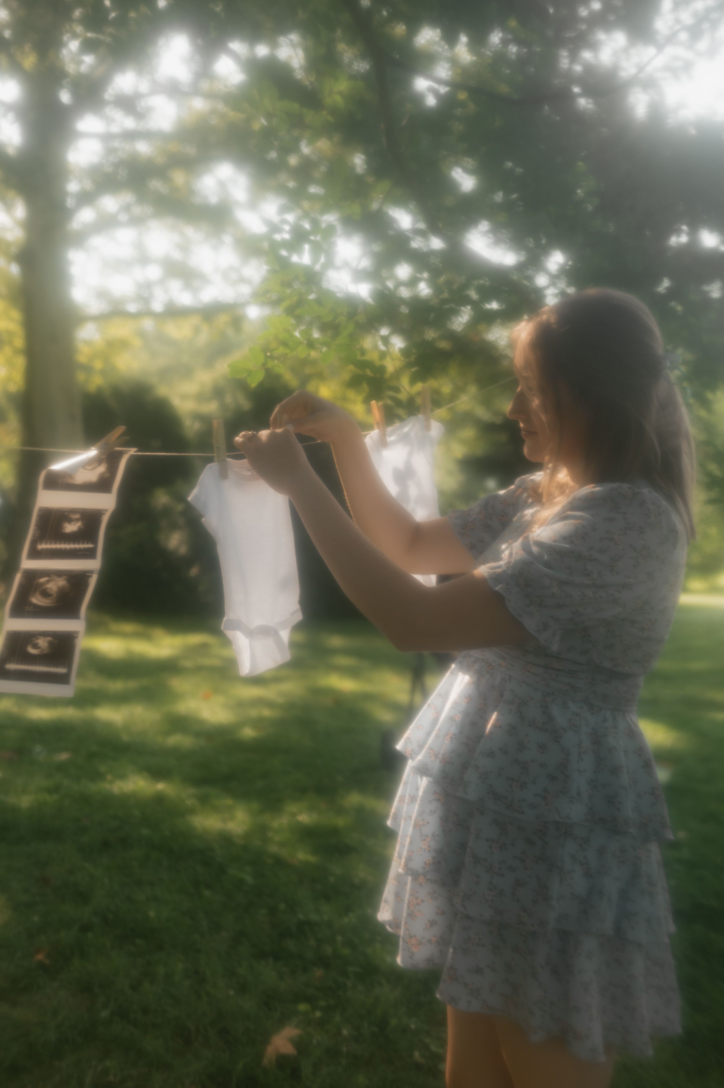 A person in a white dress hangs baby clothes on a clothesline outdoors in soft sunlit garden scenery.