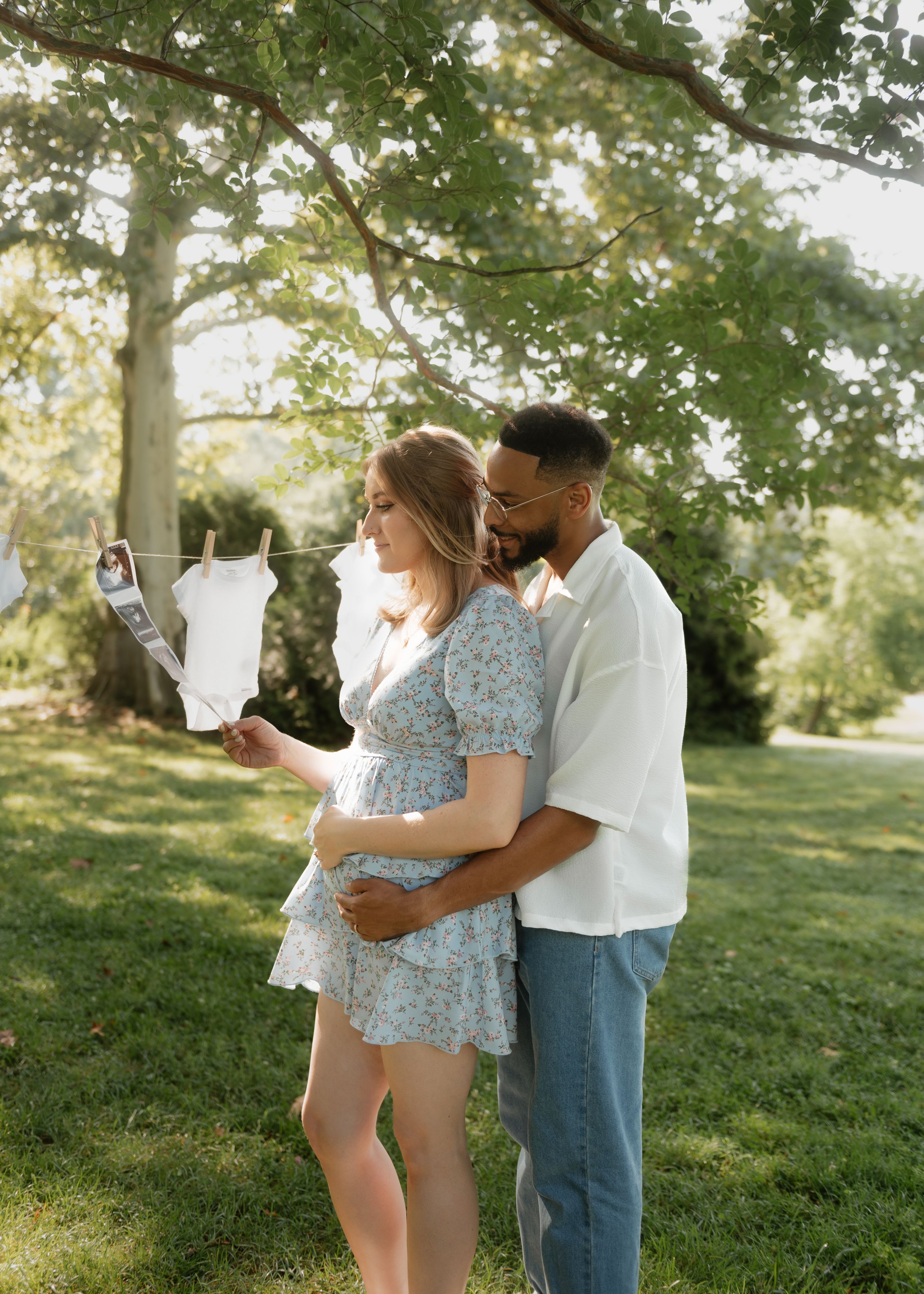 A couple in casual attire shares an intimate moment on a grassy hillside during a sunny afternoon photoshoot.