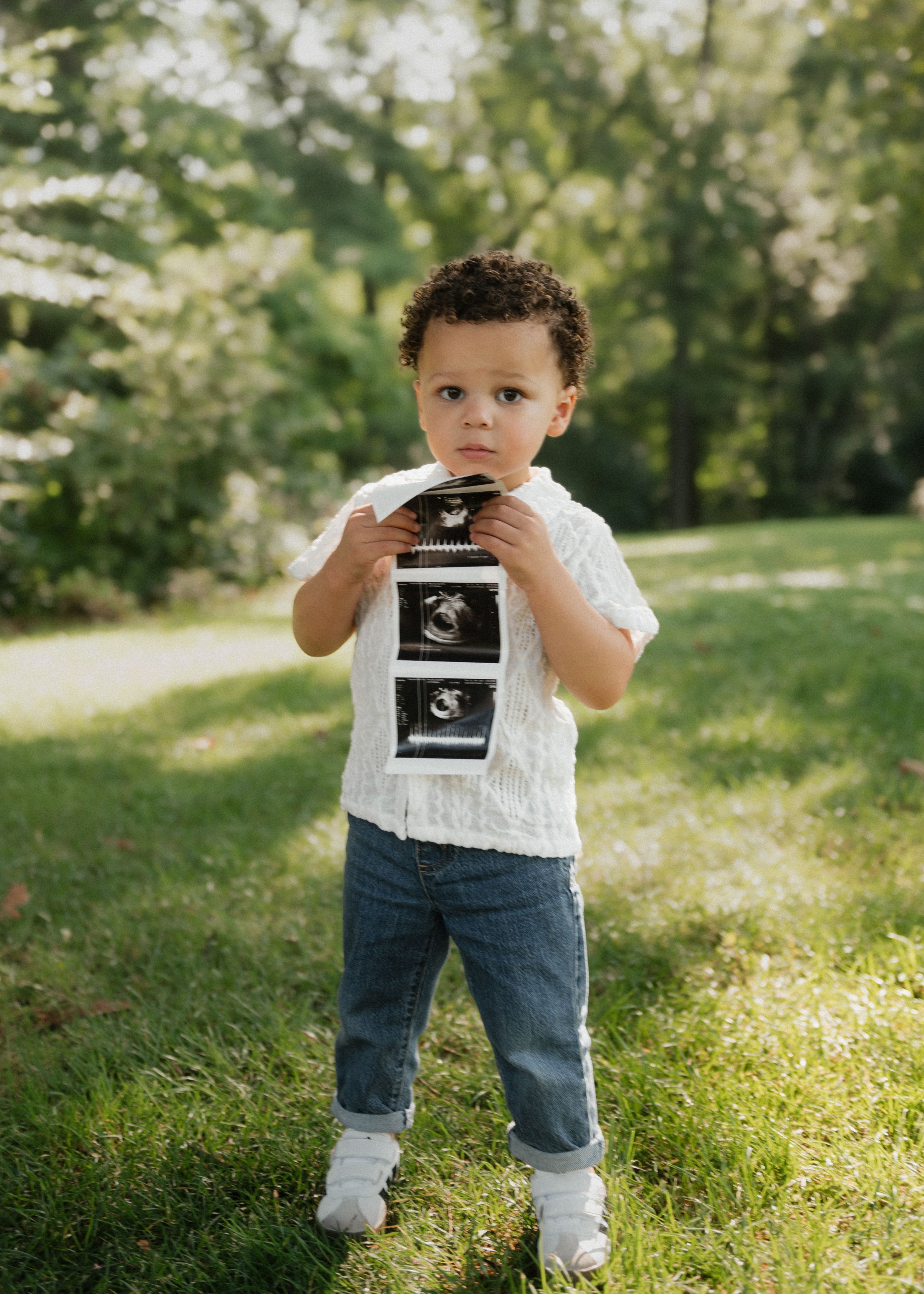 A child in blue jeans and white top stands on grass while holding ultrasound photos in multiple images.