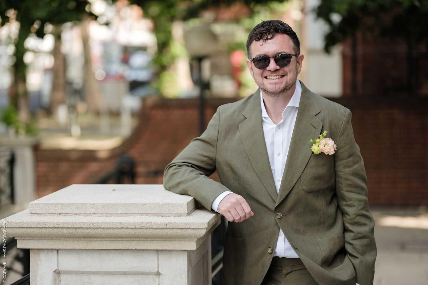 Smiling man in olive green suit with boutonniere and sunglasses leaning on stone pillar outdoors.