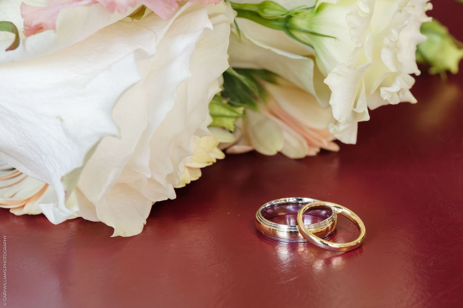 Two gold and silver wedding rings resting beside a bouquet of white roses on a red surface.