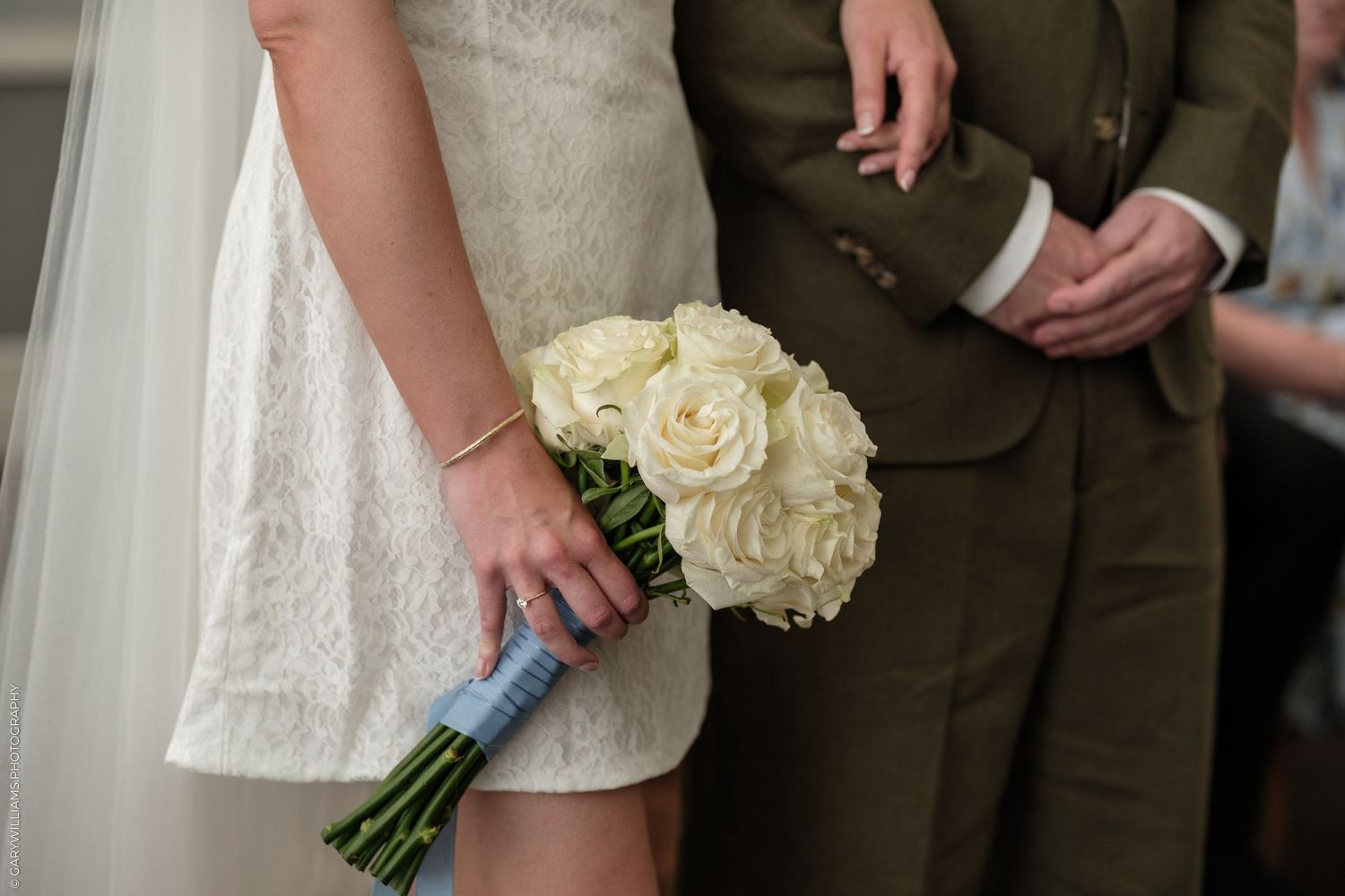 Bride in lace dress holding white rose bouquet with blue ribbon wrap, standing beside groom in olive suit.