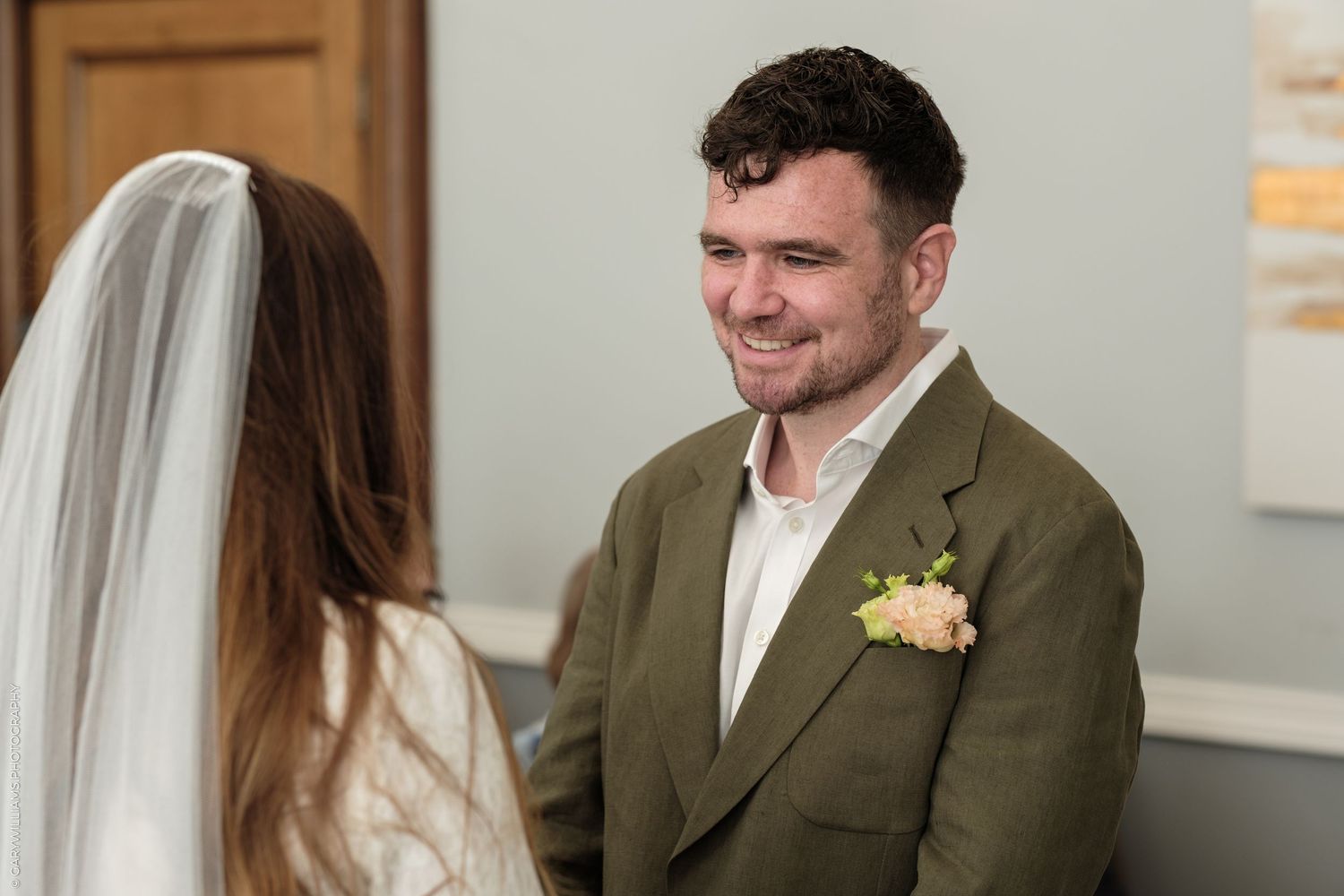Groom in olive green suit with pink boutonniere smiling at bride in white veil during wedding ceremony.