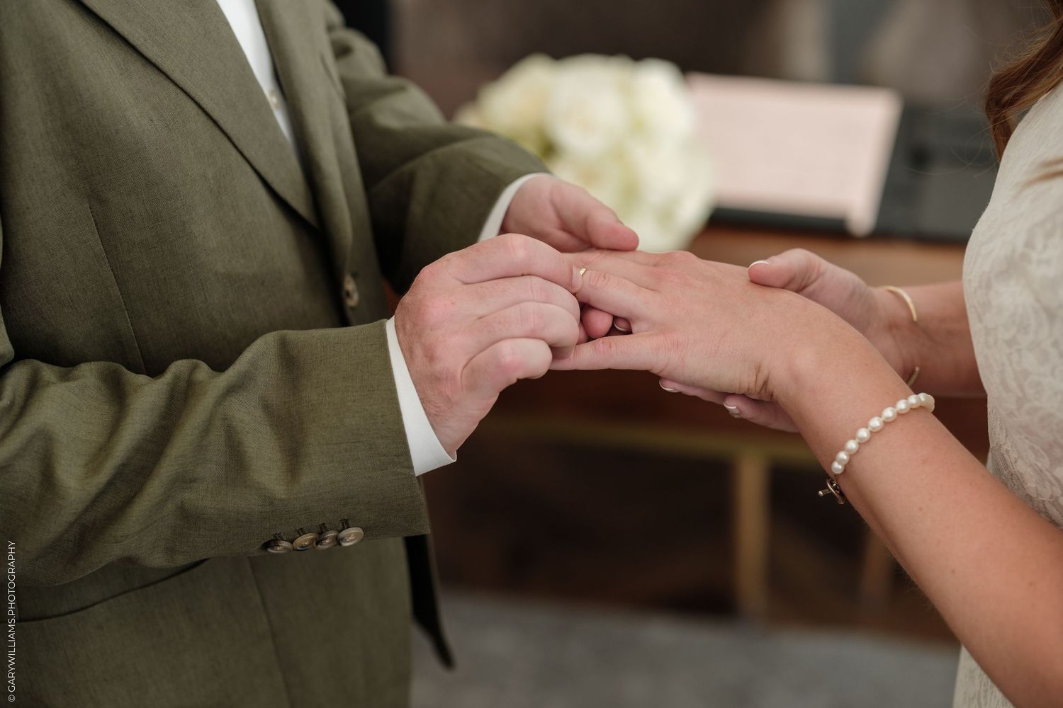 Groom placing a wedding ring on bride's finger during ceremony, bride wearing pearl bracelet.