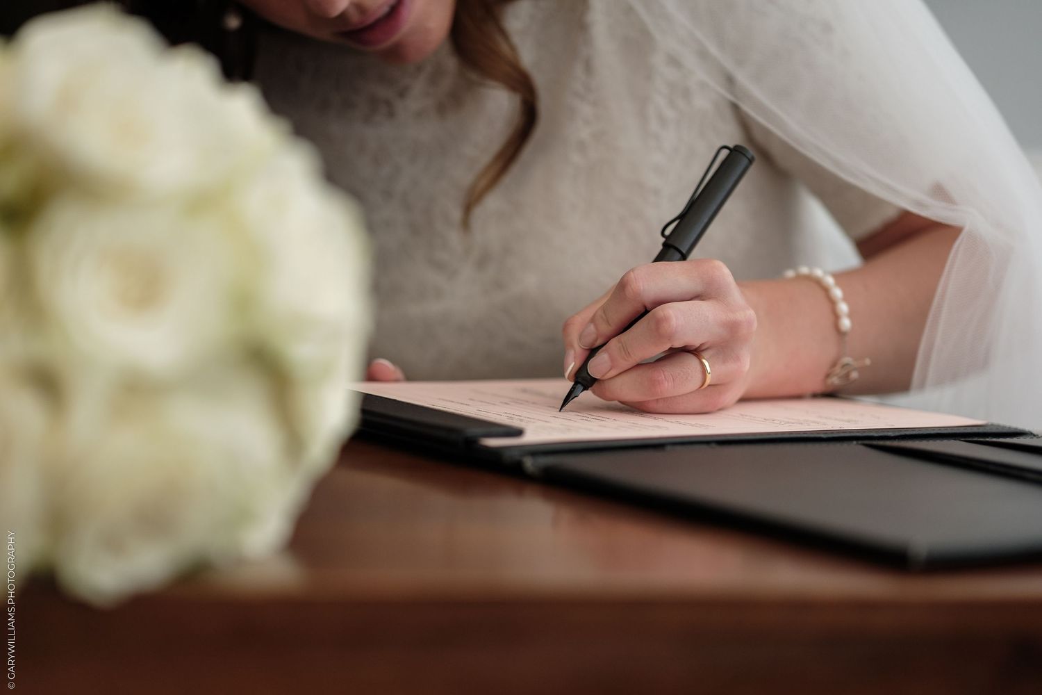 Bride signing wedding certificate with pen, wearing pearl bracelet, white bouquet in foreground.
