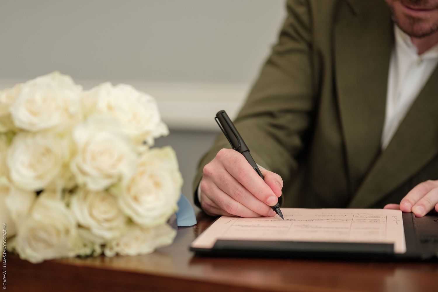 Person in green suit signing documents at desk with white roses bouquet, formal ceremony setting.