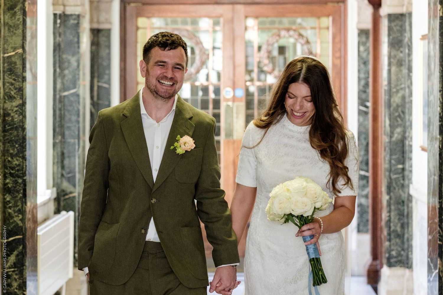Happy newlyweds walk hand-in-hand, groom in green suit with boutonniere, bride in white dress holding bouquet.